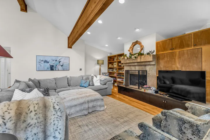 Vaulted ceiling with exposed wood beams, stone fireplace, built-in shelving, wood-finish flooring, and recessed lighting