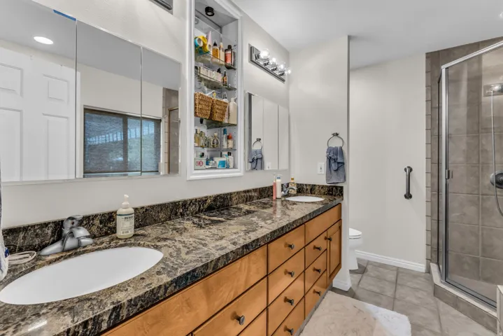 Dual vanity with stone countertop and integrated sinks, wood-finish cabinetry with multiple drawers, built-in mirrored medicine cabinets, and a glass-enclosed shower with tiled walls