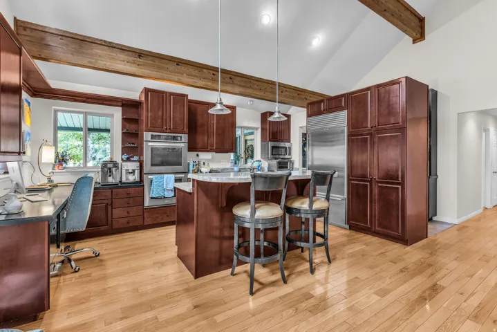 Kitchen featuring wood-finish flooring, vaulted ceilings with exposed wood beams, built-in double ovens, a central island with countertop seating, and integrated paneled cabinetry