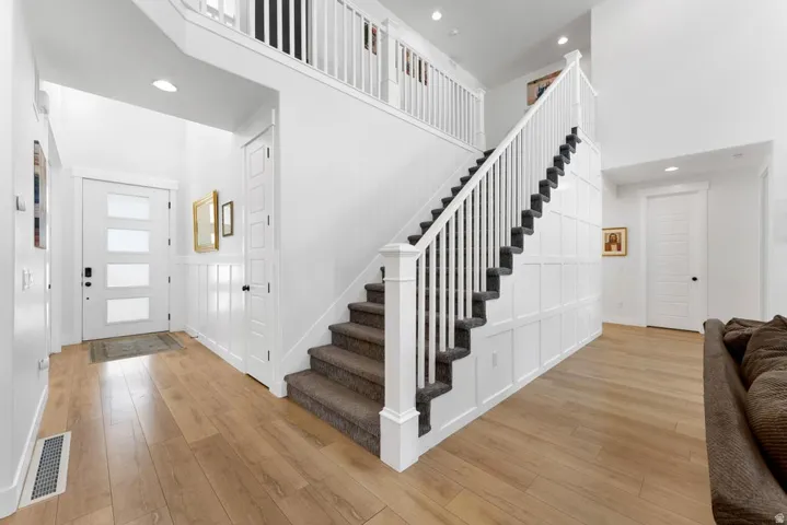 Light wood-finish flooring, a contemporary entry door with frosted panels, white paneled walls, a carpeted staircase with white balusters, and recessed ceiling lighting