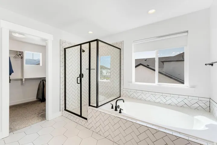 Soaking tub with black fixtures, walk-in shower with black-framed glass enclosure, white chevron-patterned tile surround, white hexagonal floor tiles, and recessed lighting