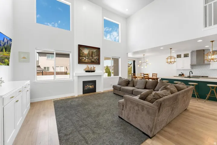Soaring ceilings featuring upper transom windows, a gas fireplace with a white mantel, light wood-finish flooring, built-in white cabinetry, and a kitchen with white cabinetry and a dark green island base