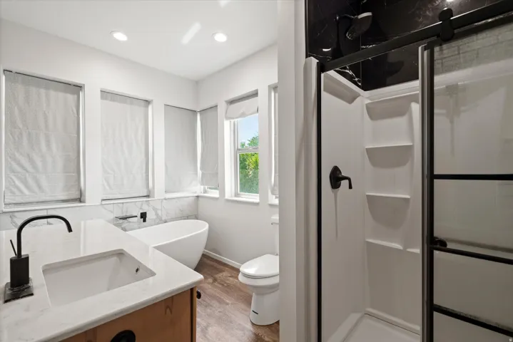 Freestanding soaking tub, walk-in shower with black-framed glass enclosure, single vanity with integrated sink, wood-finish flooring, and natural light from multiple windows
