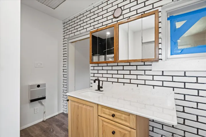 Wood-finish flooring, white brick-patterned wall, wood vanity with black hardware, white countertop with integrated sink, and dual wood-framed medicine cabinets