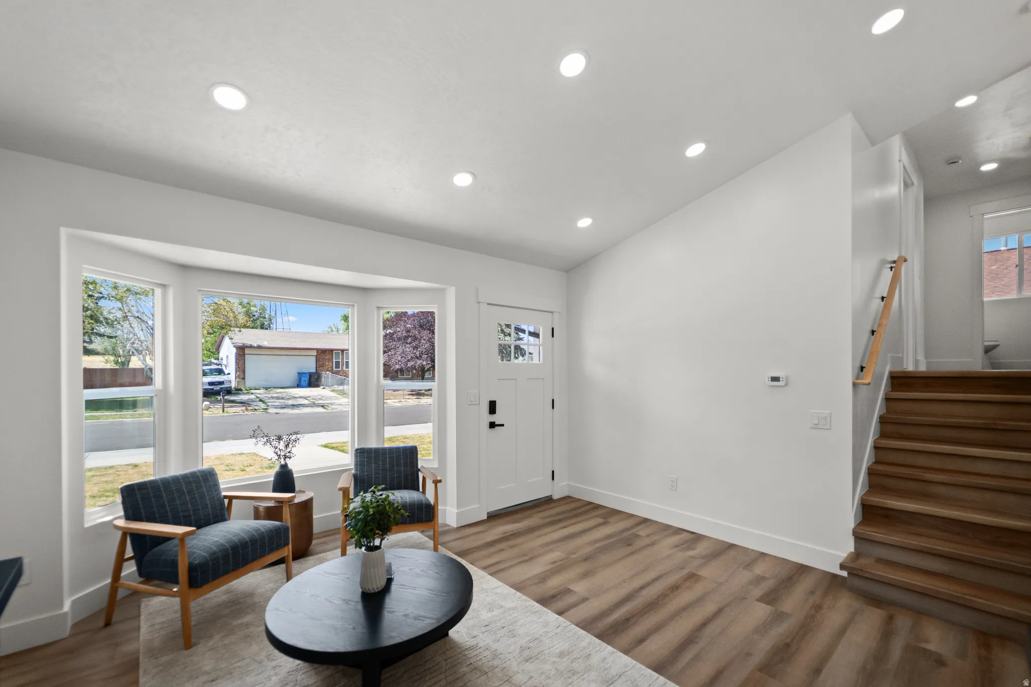 Bay window with white trim, recessed lighting, wood-finish flooring, white paneled door, and a staircase with a natural wood handrail
