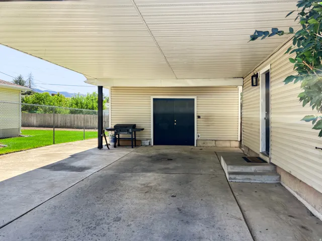 Covered carport area featuring concrete flooring, an exterior door with a wall-mounted light fixture, and beige horizontal siding