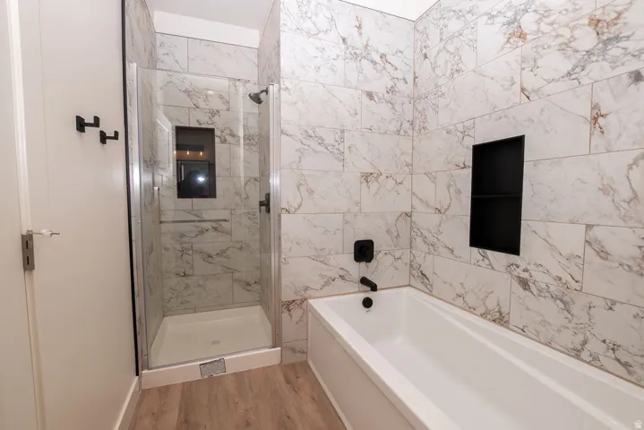 Bathroom featuring wood-finish flooring, a glass-enclosed shower, a built-in tub, and marble-look wall tiling with a recessed black niche