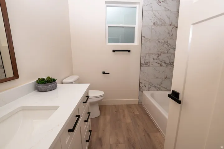 Bathroom featuring wood-finish flooring, a white vanity with black hardware, a rectangular undermount sink, a white countertop, and a shower/tub combination with tiled walls