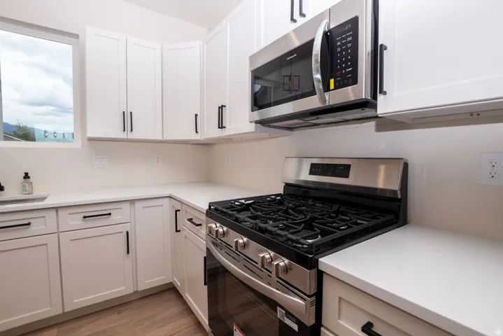 Light-toned cabinetry with matte black hardware, stainless steel gas range, coordinating stainless steel microwave, light-toned countertops, and wood-finish flooring