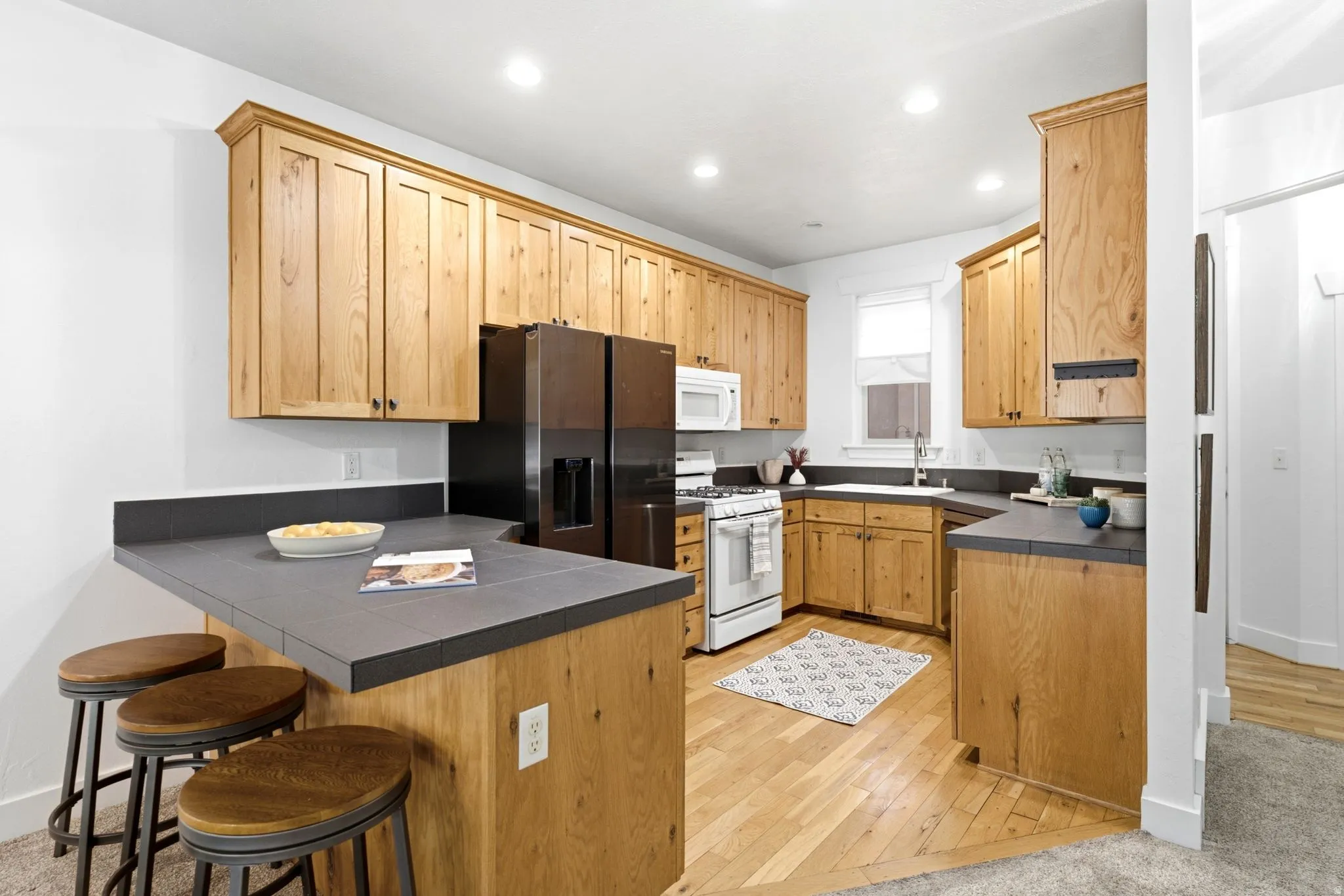 Recessed lighting illuminates the kitchen featuring wood-finish cabinetry, dark countertops, and a built-in breakfast bar