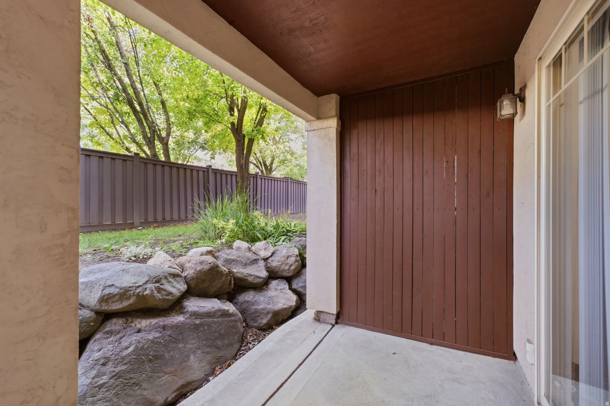 Concrete patio with a brown privacy fence, a rock garden, and mature trees beyond