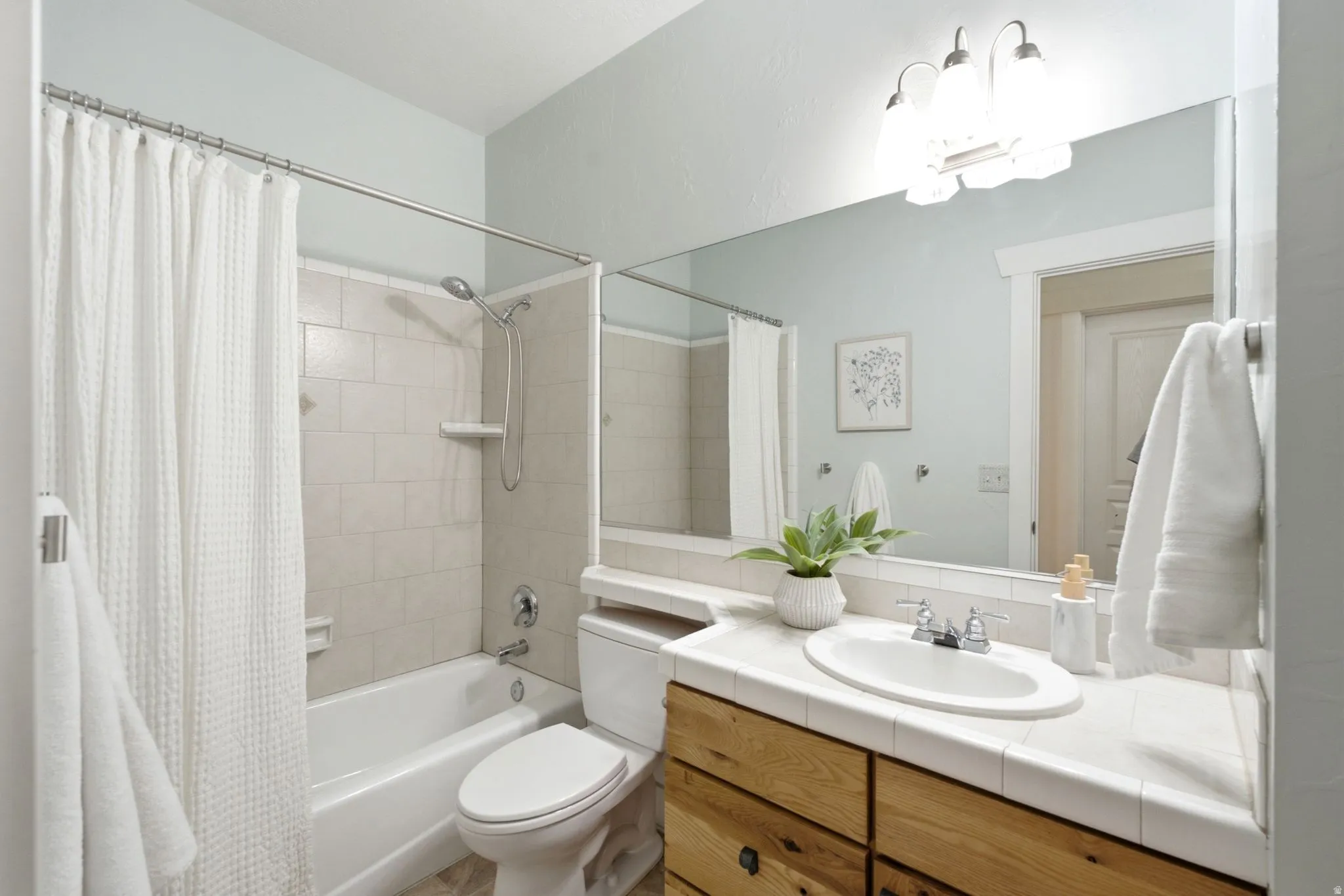 Tub-shower combination with light-toned tiled surround, wood-finish vanity with countertop sink, and a large wall-mounted mirror