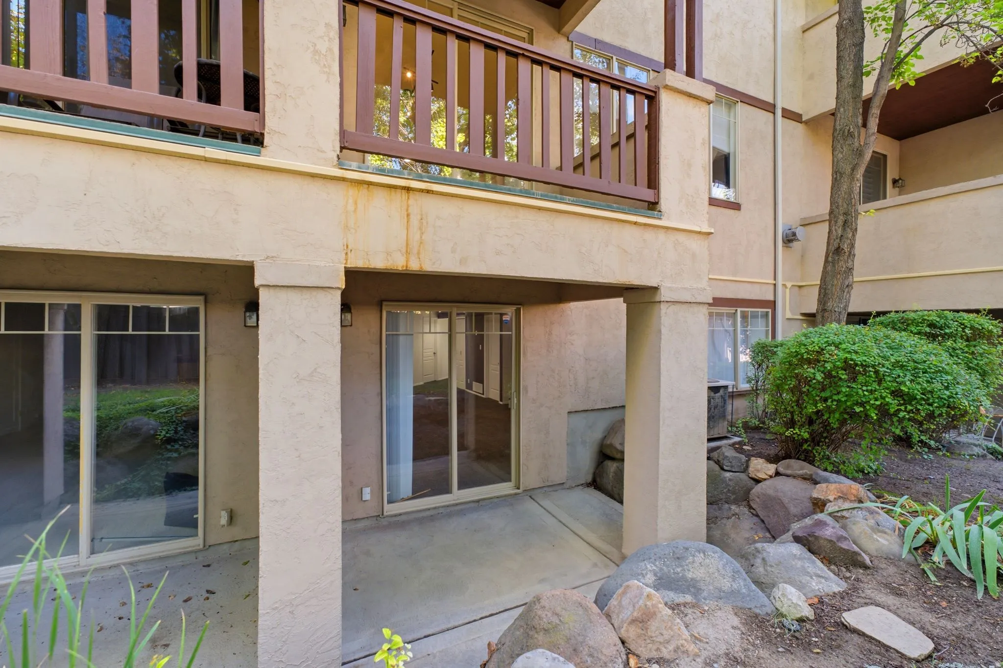 Ground-level patio with two sliding glass doors, supported by stucco-finished columns, and bordered by a natural rock landscaping feature