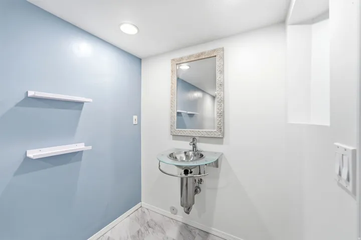 Pedestal sink with a glass basin and chrome fixtures, illuminated by a recessed ceiling light