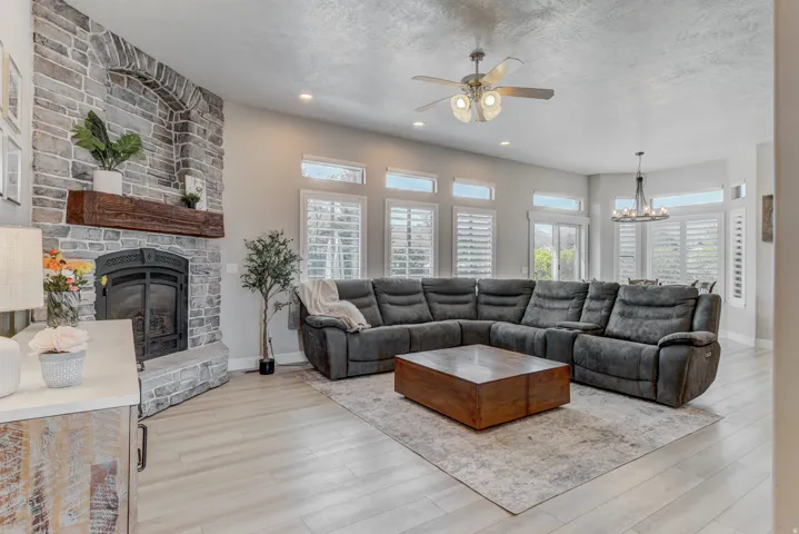 Spacious living area featuring a stone fireplace with a wood mantle, wood-finish flooring, and multiple windows with plantation shutters