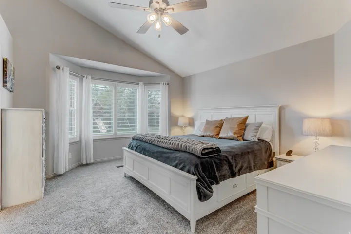Carpeted bedroom featuring a vaulted ceiling with a ceiling fan, a bay window with plantation shutters, and light gray wall tones