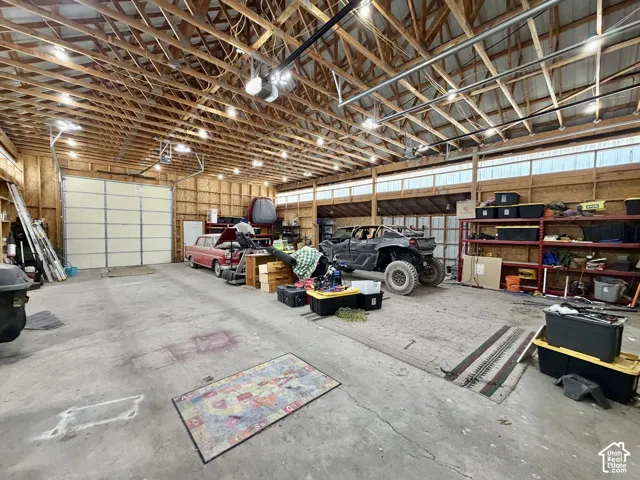 Spacious utility building featuring a concrete slab floor, exposed wood beam ceiling, multiple overhead light fixtures, extensive shelving, and a large roll-up door