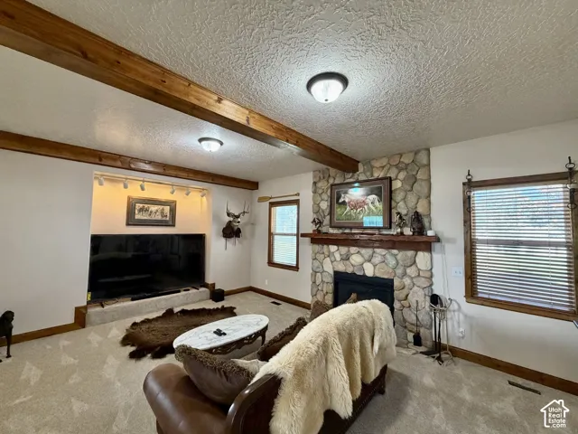 Exposed wood beams, stone-clad fireplace with wood mantle, carpeted flooring, built-in media niche with track lighting, and wood-framed windows