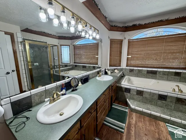 Dual vanity with integrated sinks, full-width mirror with overhead lighting, wood-finish flooring, tiled soaking tub, and a separate glass-enclosed shower