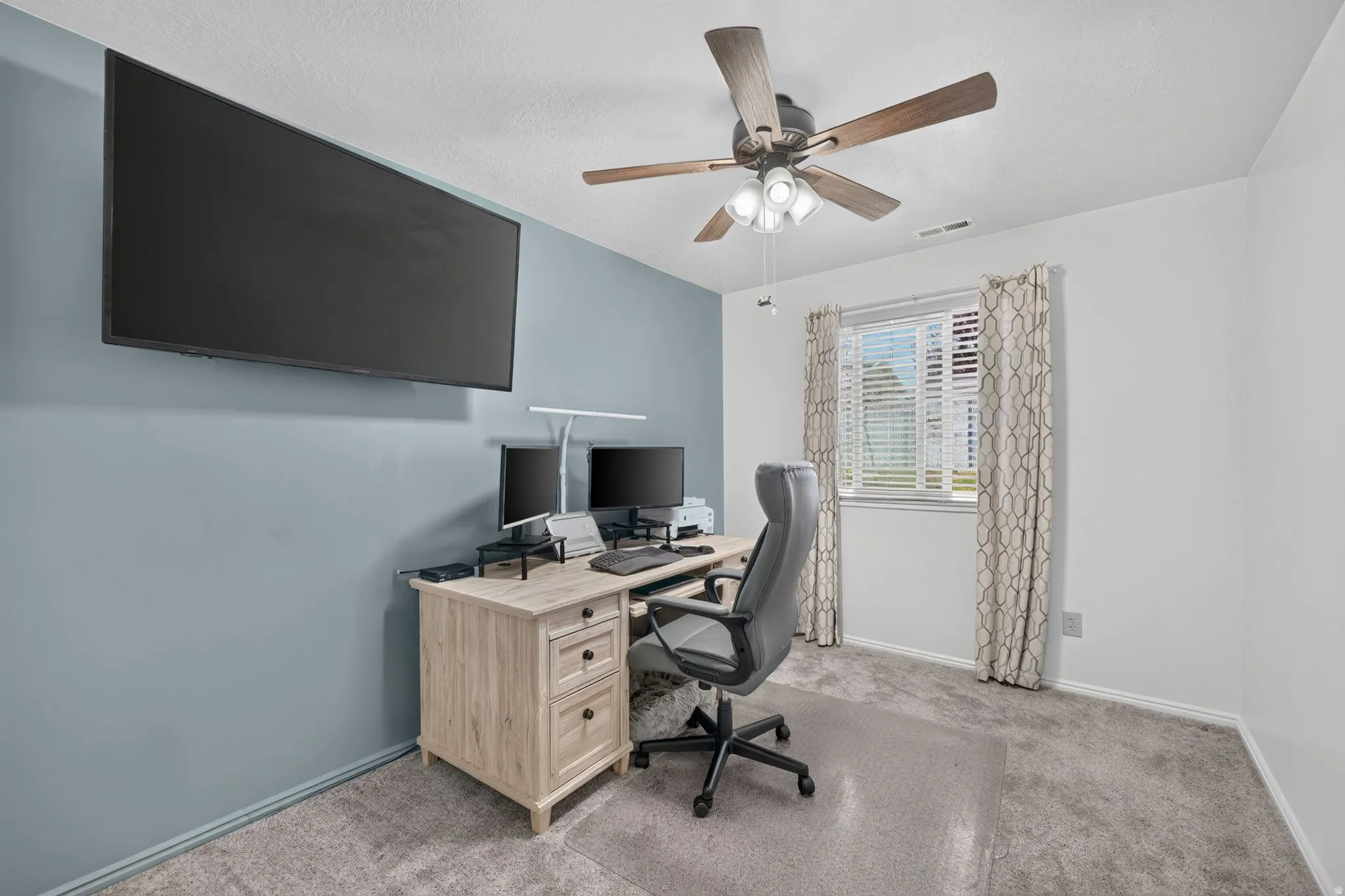 Modern home office featuring a light wood desk with storage drawers, ergonomic black task chair, and dual monitor setup