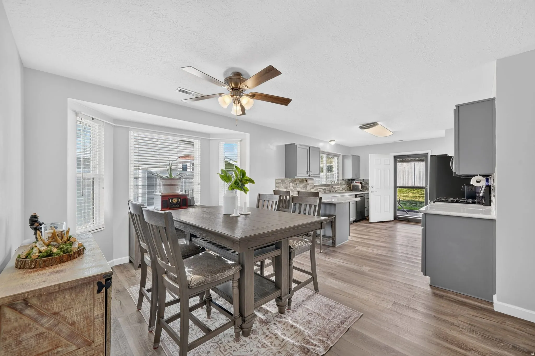 Open-concept living and dining area featuring wood-finish flooring, a ceiling fan with brushed metal accents, and abundant natural light from multi-pane windows