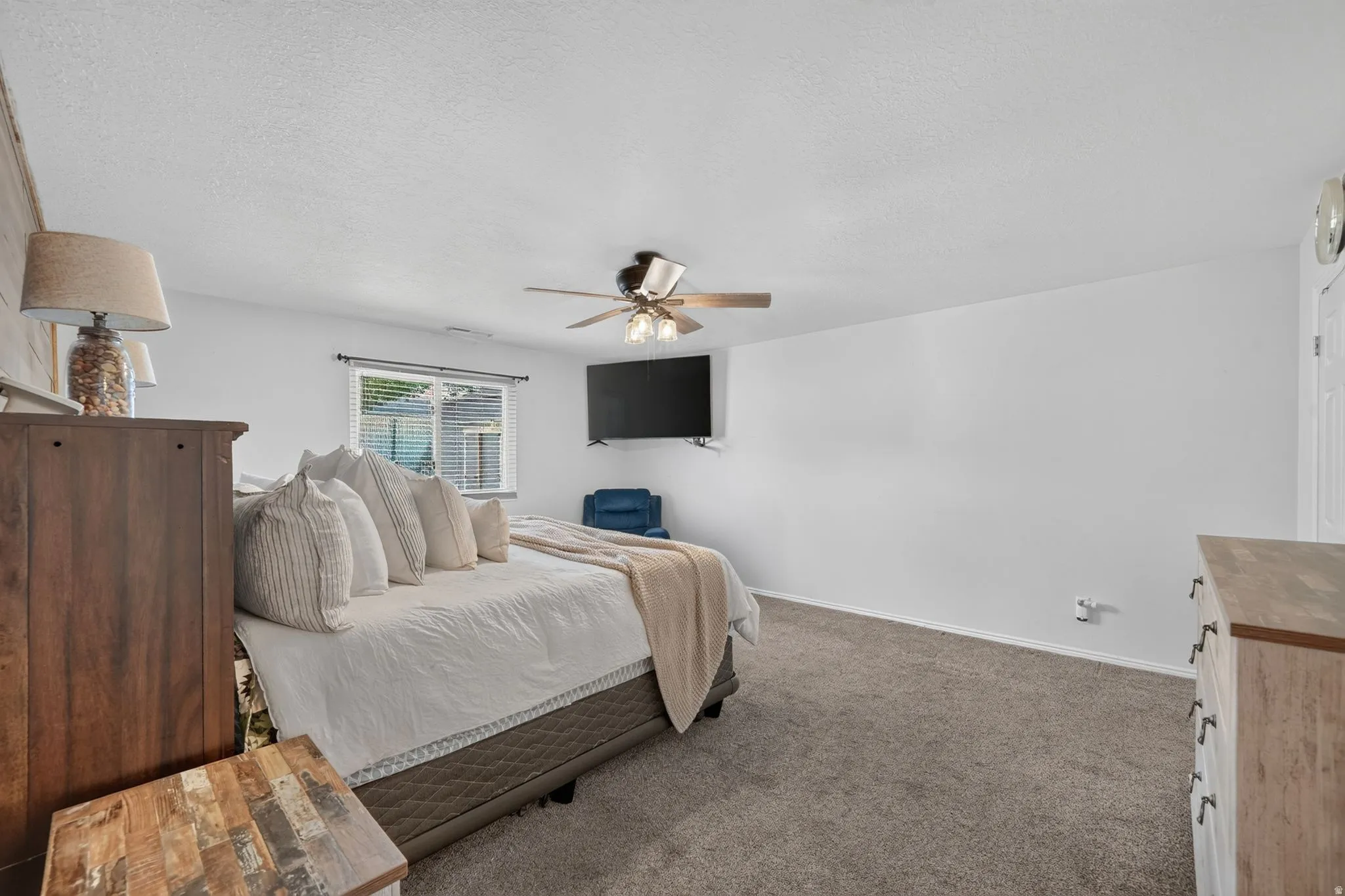 Spacious primary bedroom featuring neutral décor, carpeted flooring, ceiling fan with integrated lighting, wall-mounted flat-screen television, and abundant natural light from multiple windows