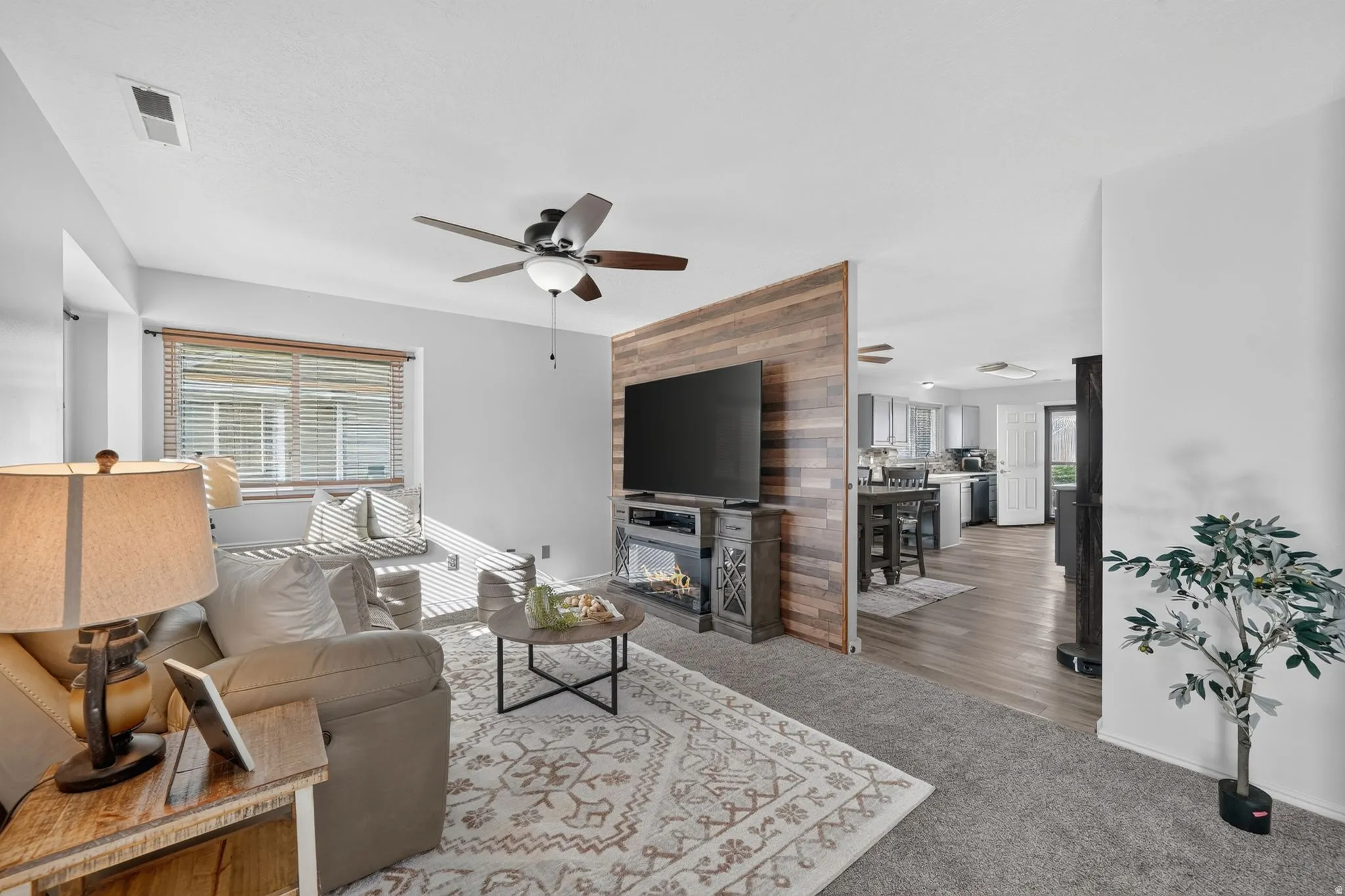 Wood-finish accent wall with an electric fireplace, ceiling fan with integrated lighting, window with horizontal blinds, and wood-finish flooring extending into the kitchen