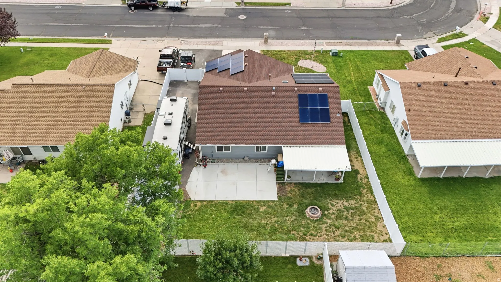 Aerial view showcasing a well-appointed residential property featuring an asphalt shingle roof, attached garage with driveway access, expansive manicured lawn with mature landscaping, and solar panel installation on the roof