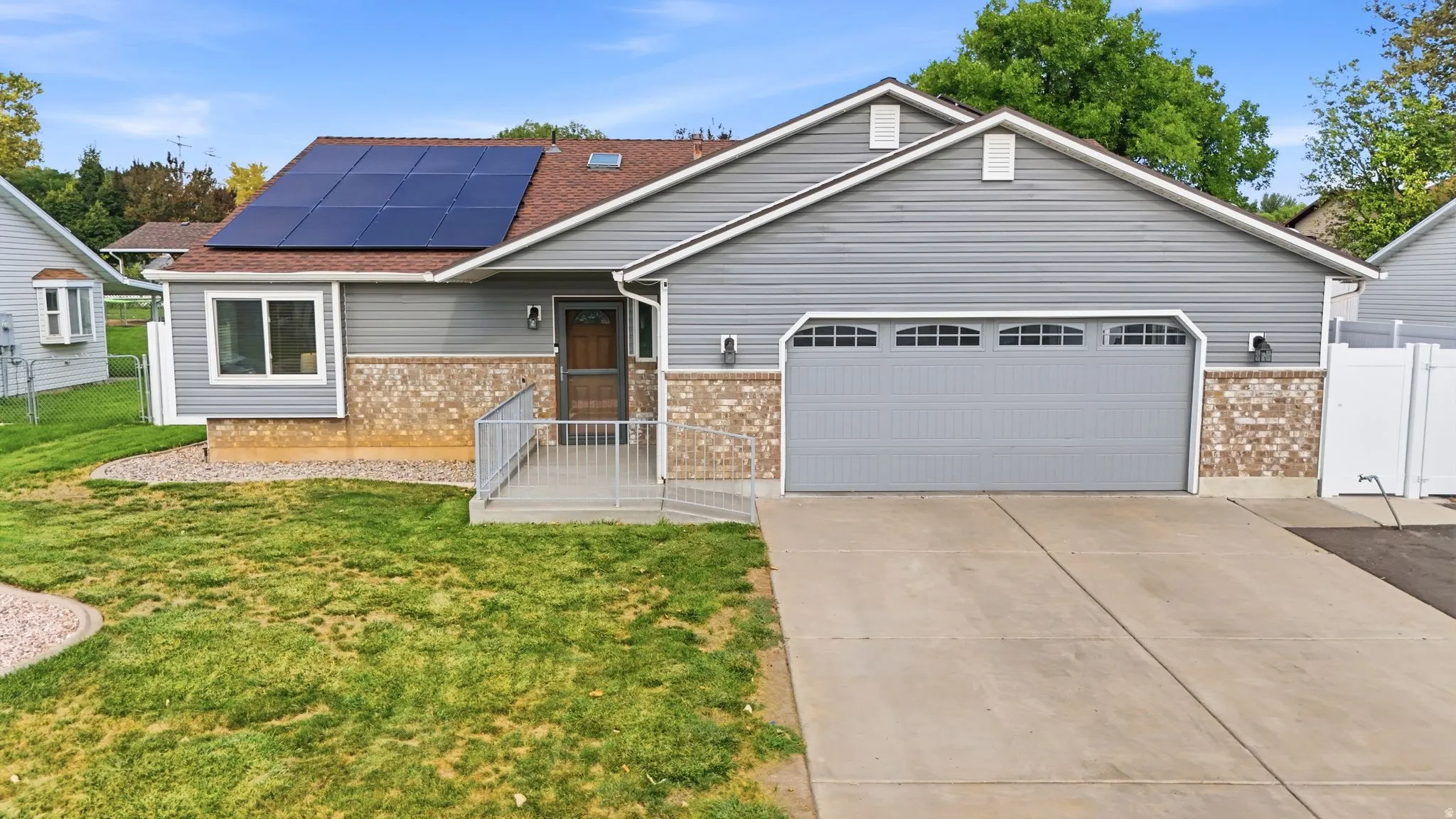 Modern ranch-style home featuring a two-car garage with paneled doors, stone and siding exterior, a covered front porch with composite decking, solar panel array on the roof, and a level concrete driveway