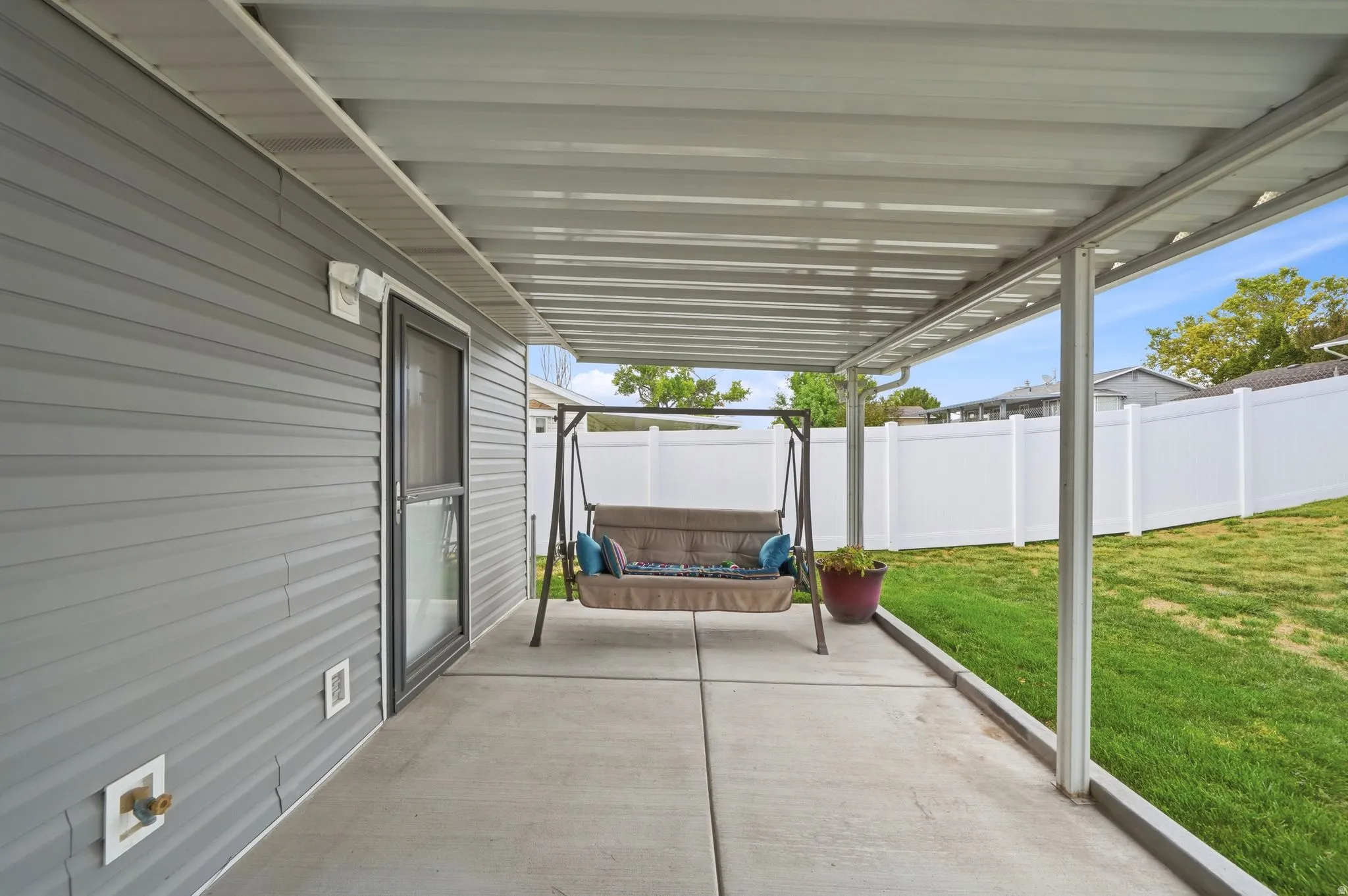 Covered patio with concrete flooring, framed by a white vinyl privacy fence and a manicured lawn