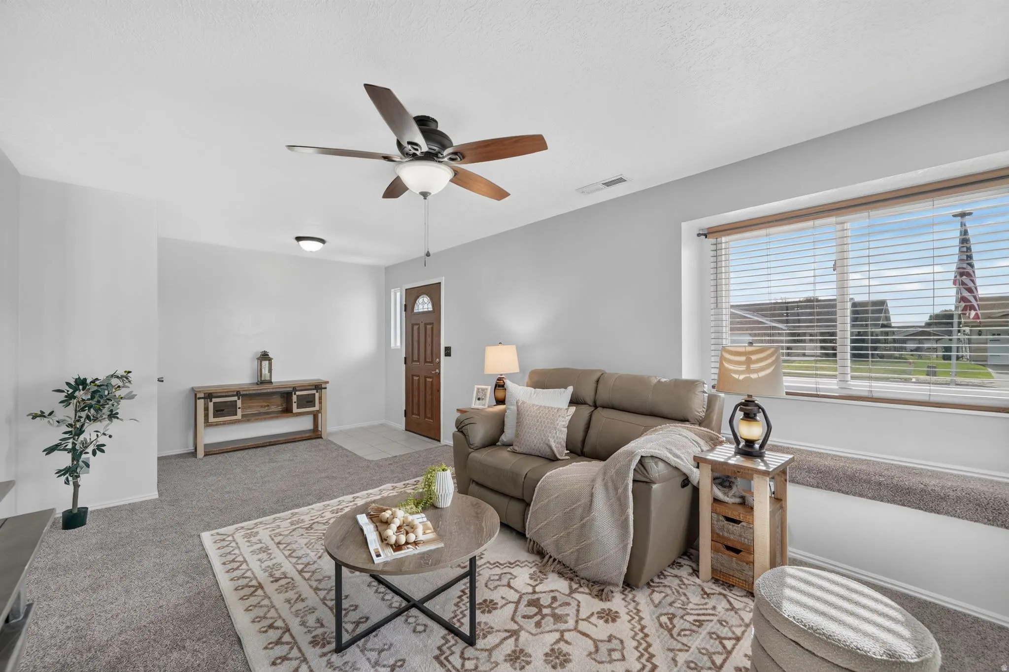 Bright living room showcasing neutral carpeting, a ceiling fan with integrated lighting, and expansive windows with blinds