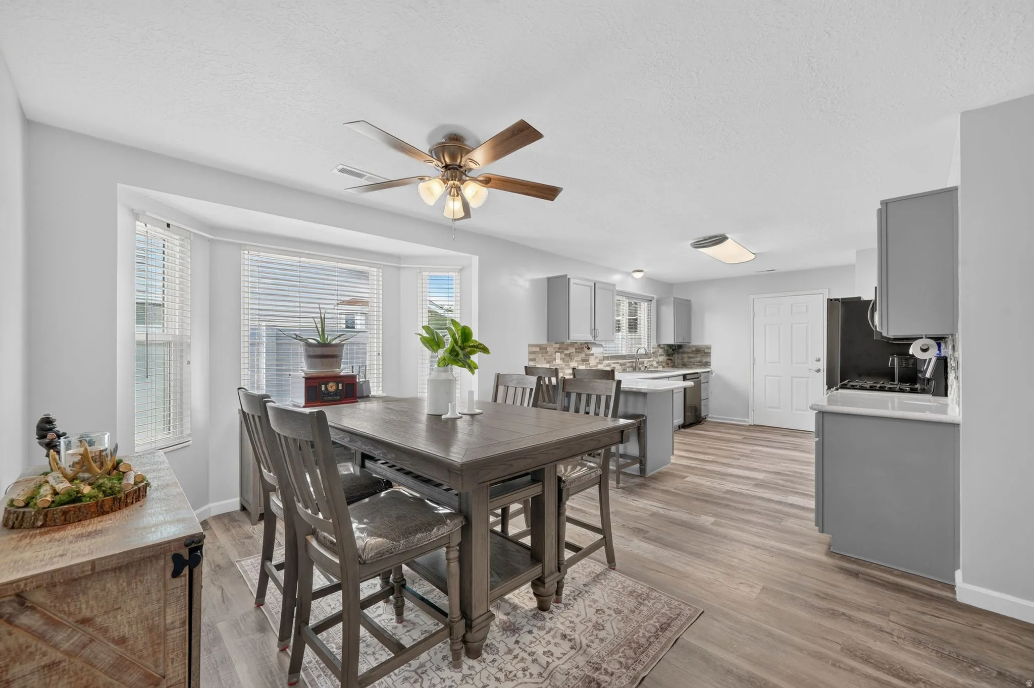 Bay window with blinds, wood-finish flooring, ceiling fan with light fixture, gray cabinetry, and tile backsplash