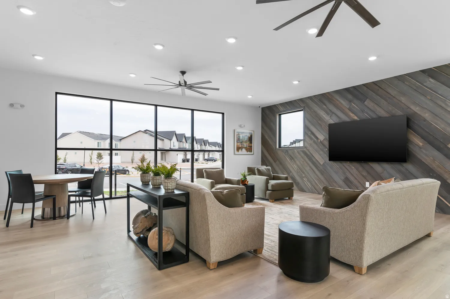Expansive windows with black framing, wood-finish flooring, recessed lighting, and a diagonal wood-paneled accent wall with a mounted television