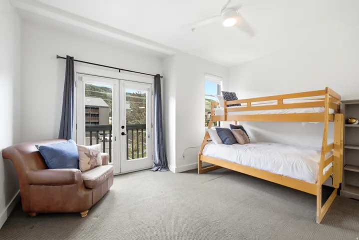 White walls, gray carpeted flooring, a ceiling fan, a window, and double doors opening to a balcony with a dark railing