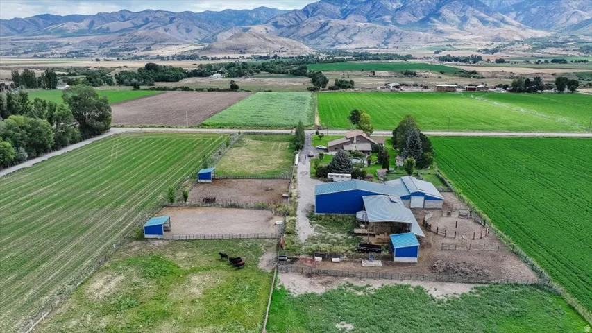 Expansive agricultural property featuring multiple blue-roofed outbuildings, fenced animal enclosures, and cultivated fields with mountain scenery
