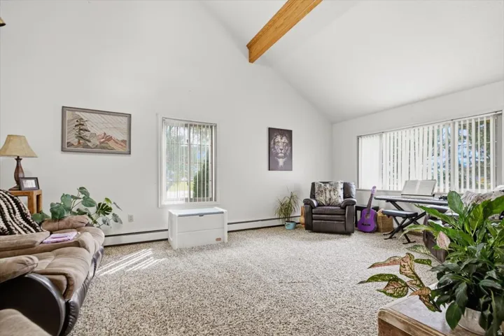 Vaulted ceiling with exposed wood beam, large windows with vertical blinds, and wall-to-wall carpeting