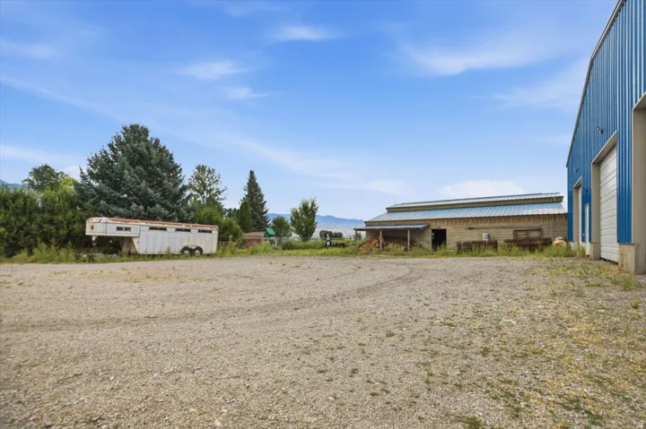 Gravel lot with a blue metal-sided building featuring two roll-up doors, a brick-sided building with a covered porch, and mature trees
