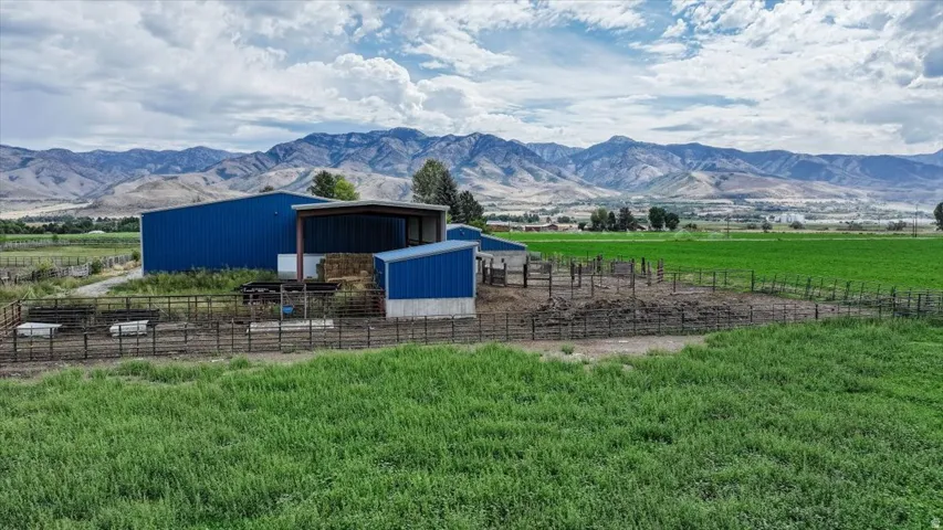 Expansive green fields with mountain backdrop, featuring a large blue metal agricultural building, an open-sided shed, and fenced outdoor enclosures