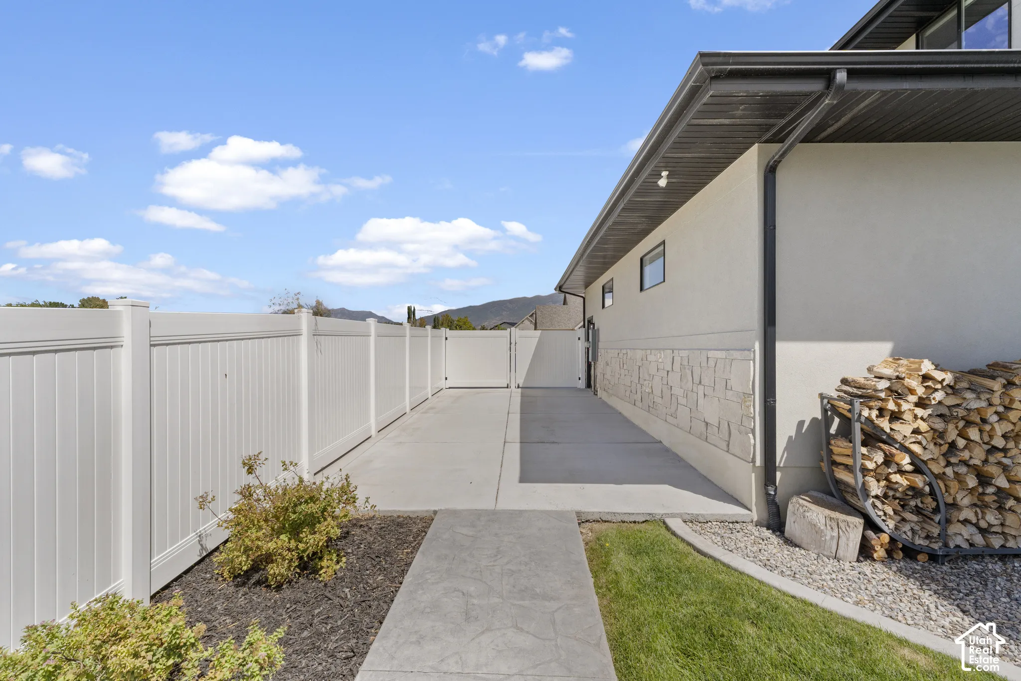 Expansive rear courtyard featuring a concrete pathway, composite fencing, manicured lawn with mulched garden beds, and stacked firewood storage