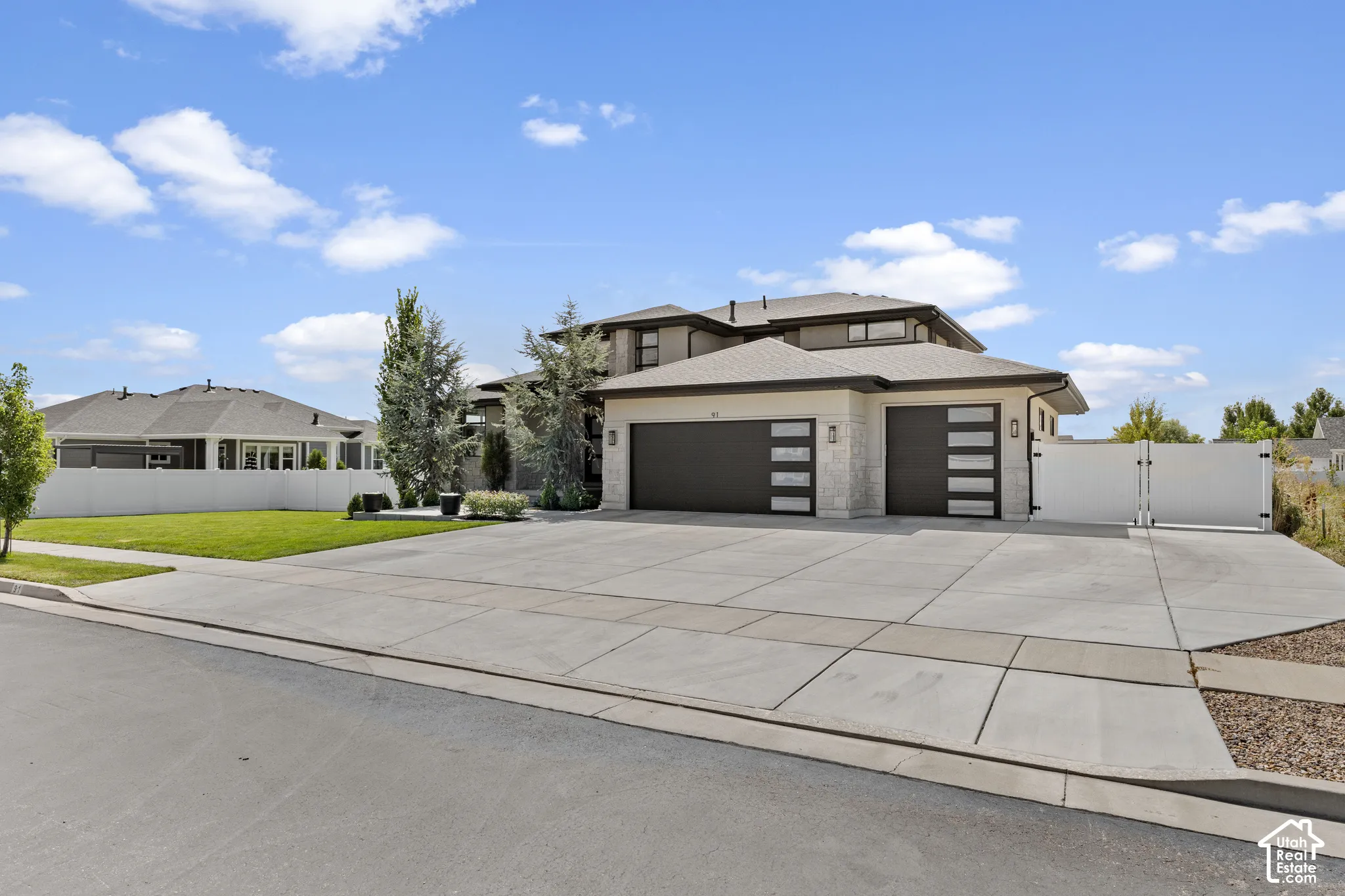 Modern two-story residence featuring a substantial concrete driveway with dual three-car garage bays, dark-finish garage doors with horizontal paneling, and mature landscaping including evergreen trees and manicured lawn