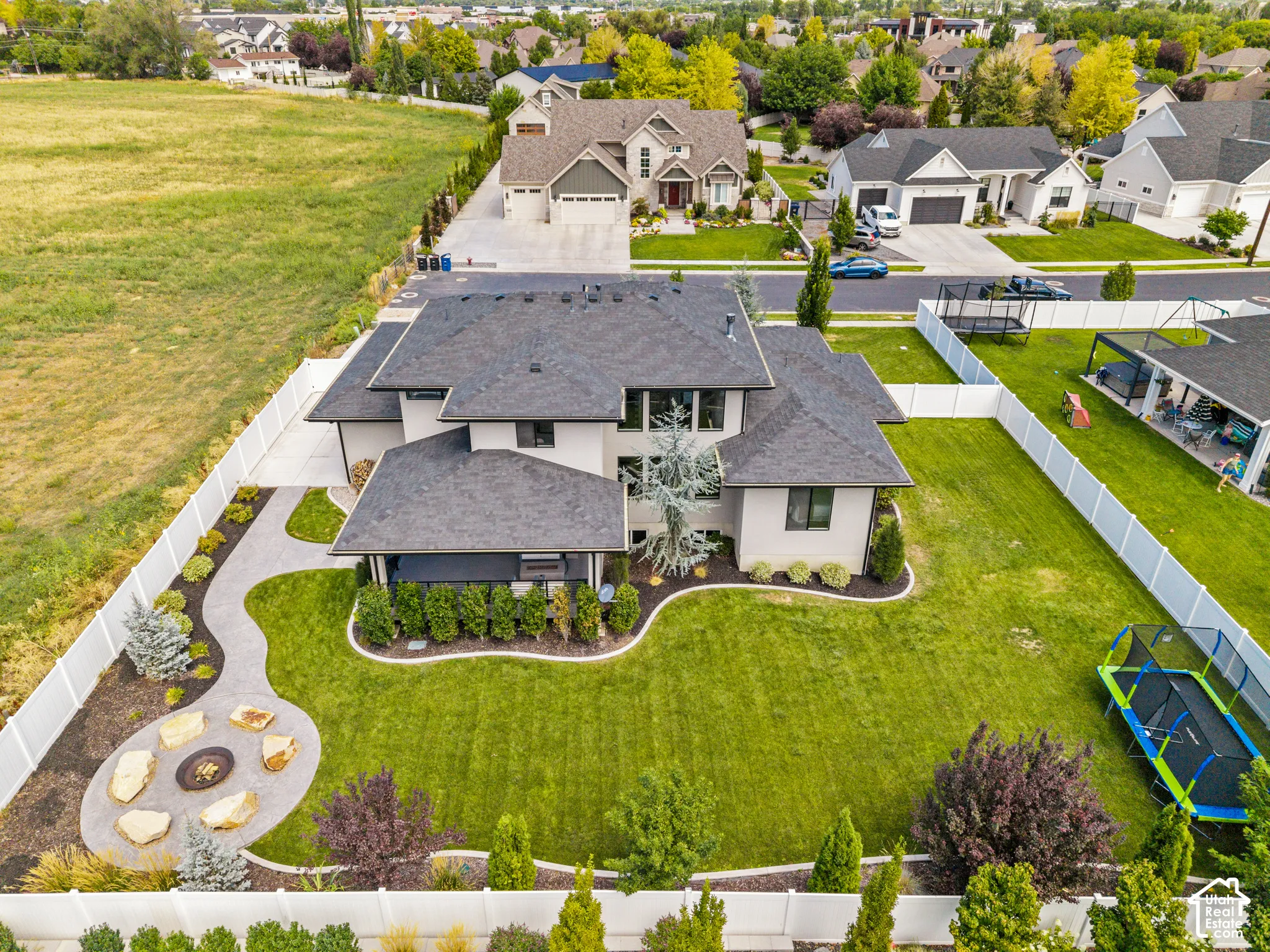 Stunning modern estate showcasing a manicured lawn with mature landscaping, circular hardscaped patio with fire pit feature, and a two-story residence with dark asphalt shingle roofing and light-toned exterior