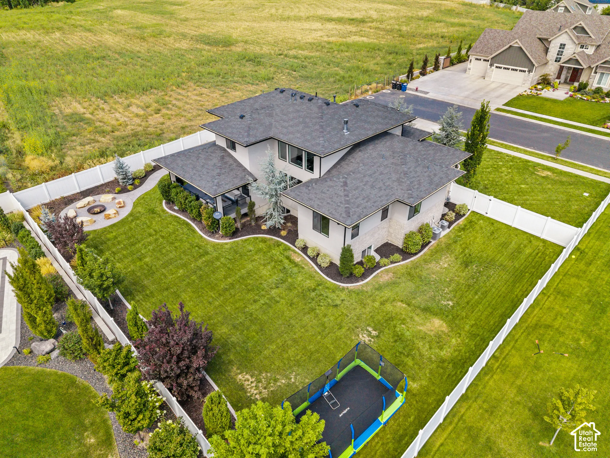 Stunning contemporary residence showcasing a curved architectural design with charcoal asphalt shingle roofing, set on expansive manicured grounds featuring mature landscaping, defined perimeter hardscaping, and a recreational trampoline installation
