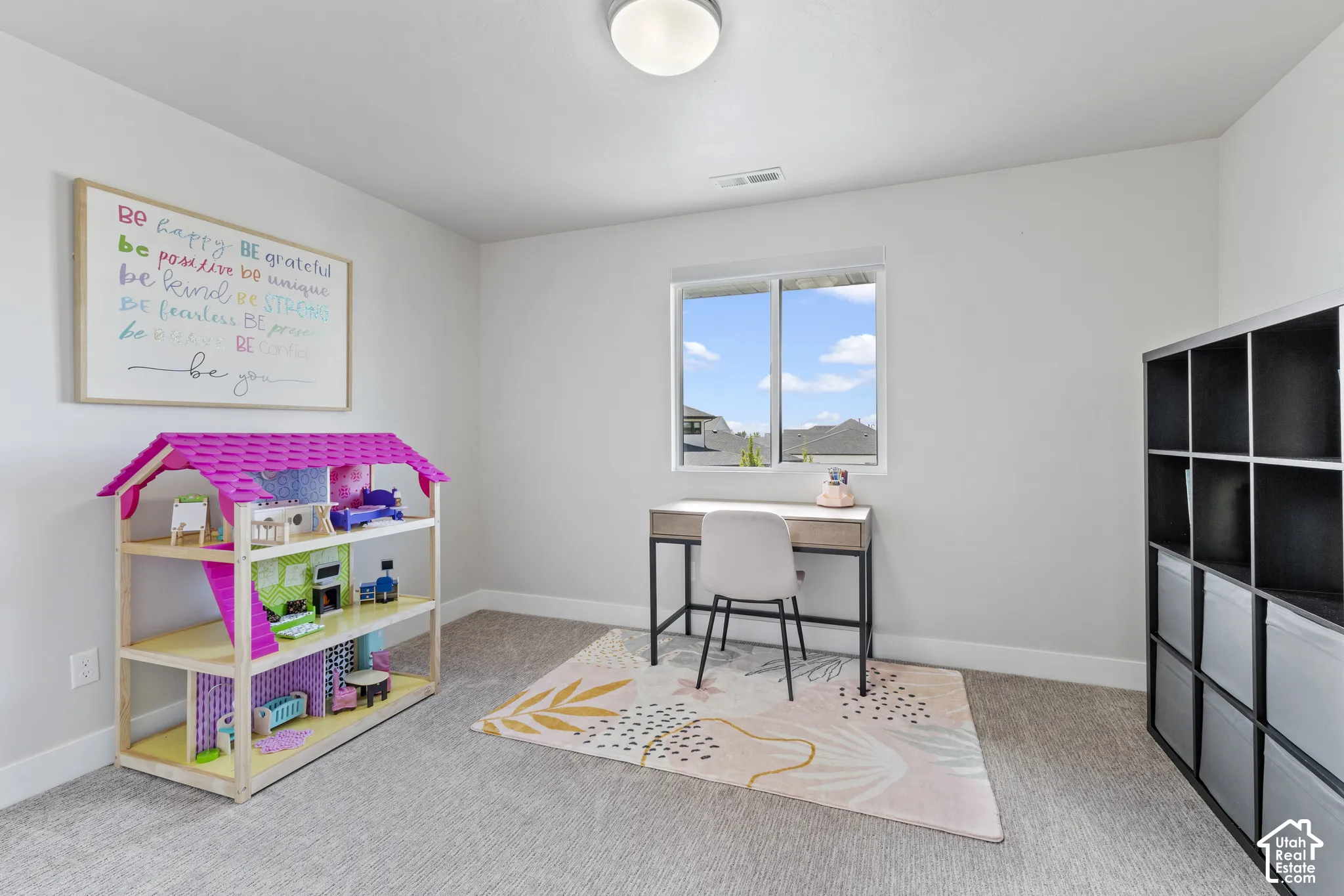 Bright, multi-purpose room featuring large windows with natural light, neutral carpeting, built-in black storage cubbies, and a dedicated work area with desk and chair