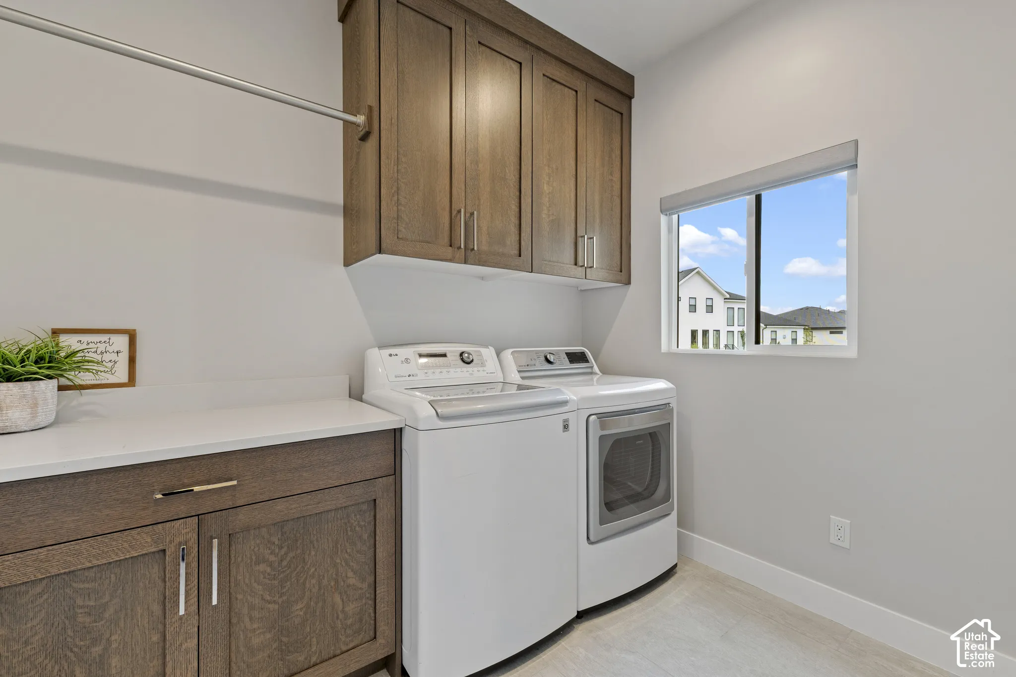 Dedicated laundry room featuring white front-load washer and dryer, wood-finish cabinetry with ample storage, quartz countertop workspace, and natural light from a large picture window overlooking the neighborhood