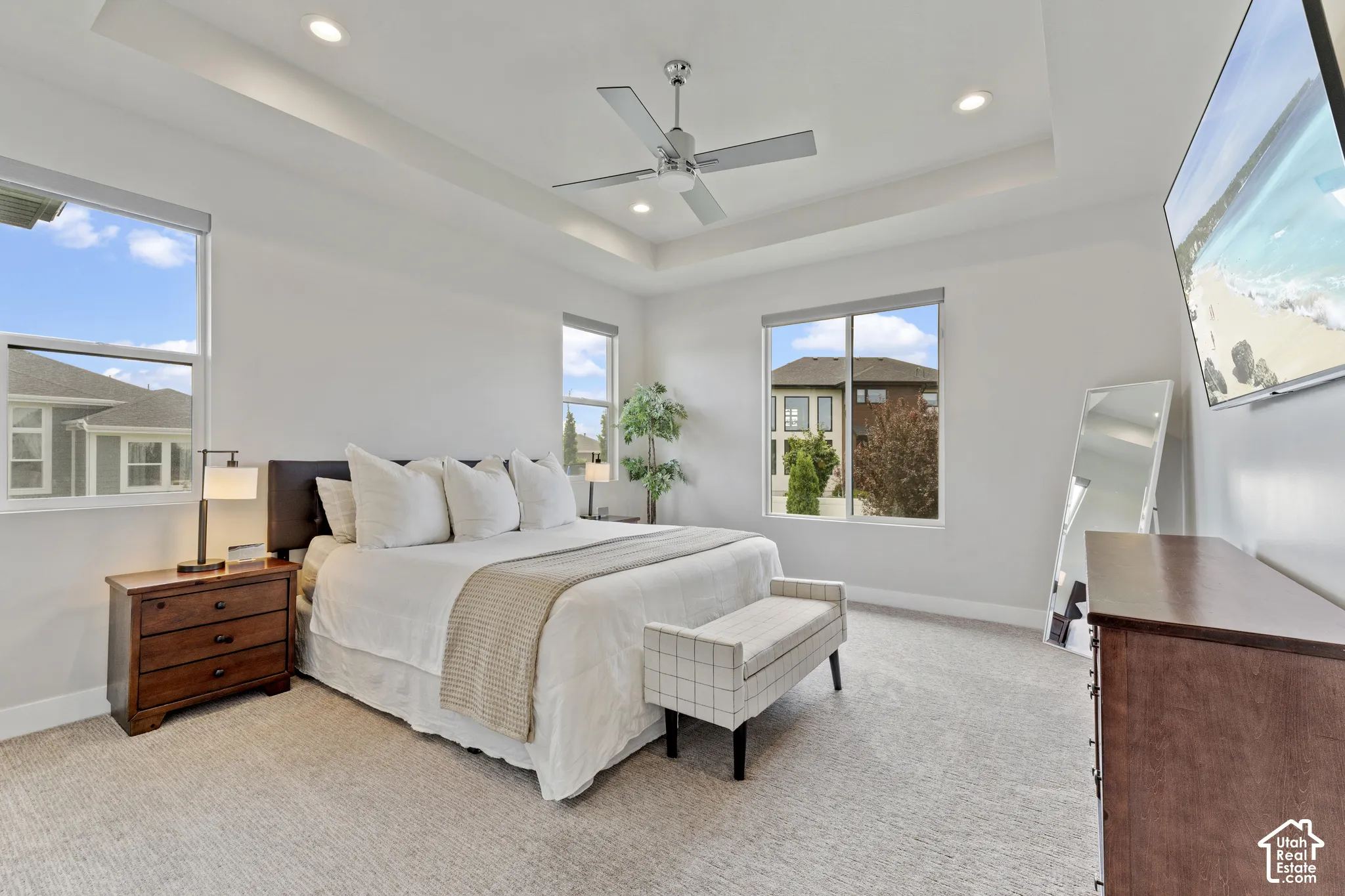 Bright master bedroom showcasing multiple windows with natural light, ceiling fan with recessed lighting, neutral carpet flooring, and wood-finish nightstand