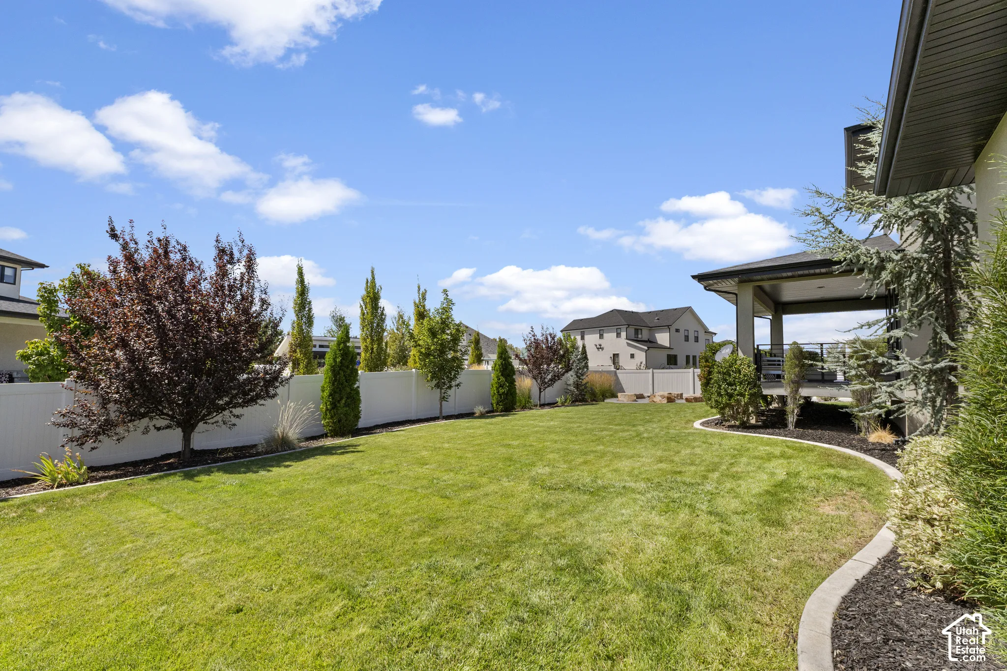 Expansive backyard featuring a manicured lawn, mature landscaping with evergreen and deciduous trees, concrete hardscaping with curved edging, and a covered patio structure