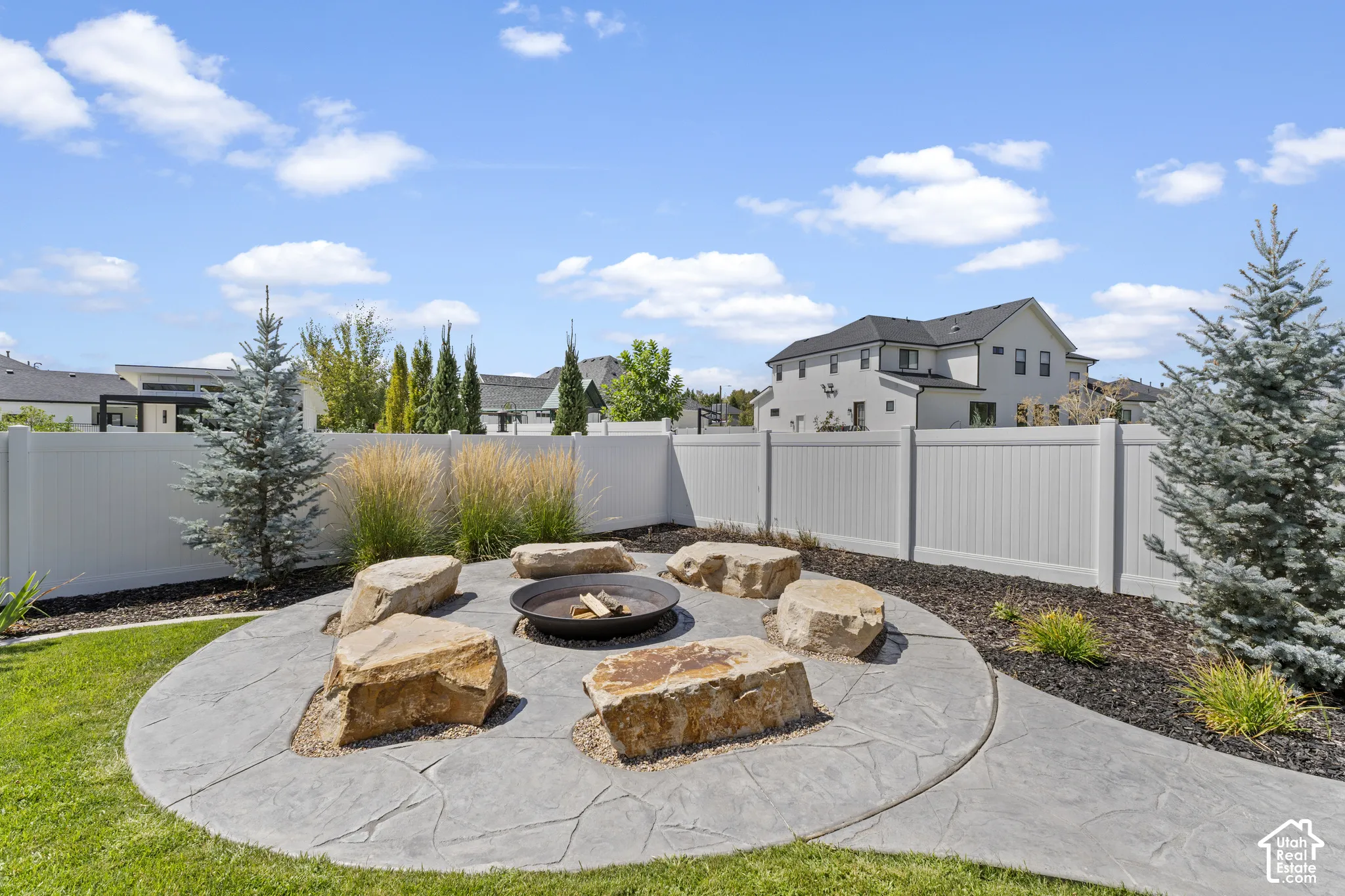 Expansive backyard oasis featuring a circular stamped concrete patio with integrated fire pit, surrounded by mature landscaping including ornamental grasses and evergreen plantings