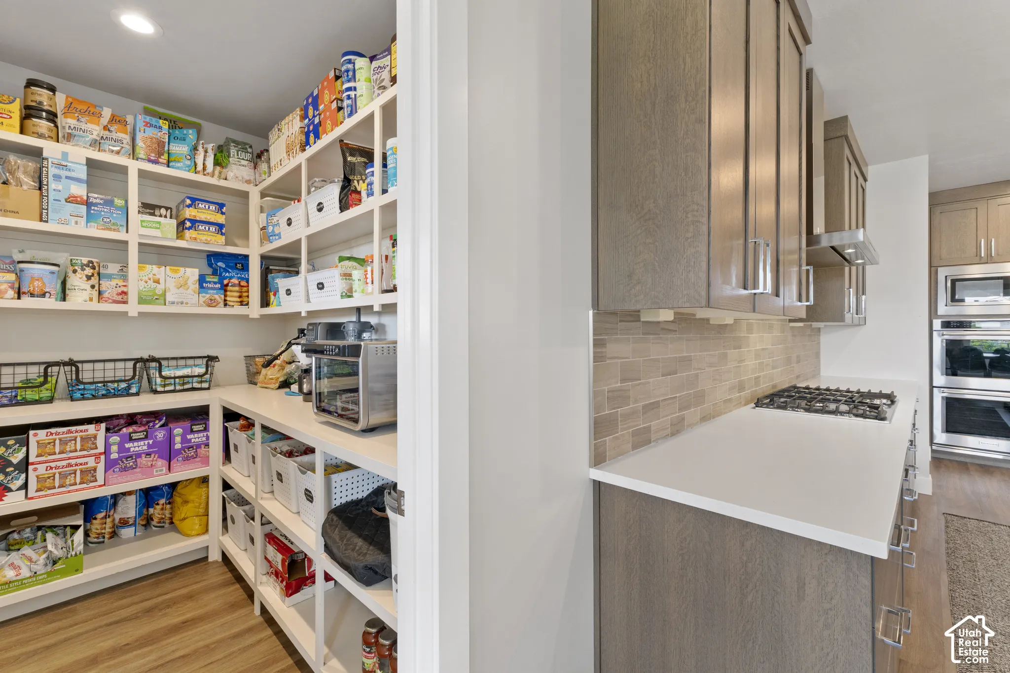 Generous walk-in pantry featuring floor-to-ceiling shelving with pull-out drawers, ample storage for dry goods and small appliances, wood-finish flooring, and direct access to the kitchen with quartz countertops and stainless steel gas cooktop visible bey