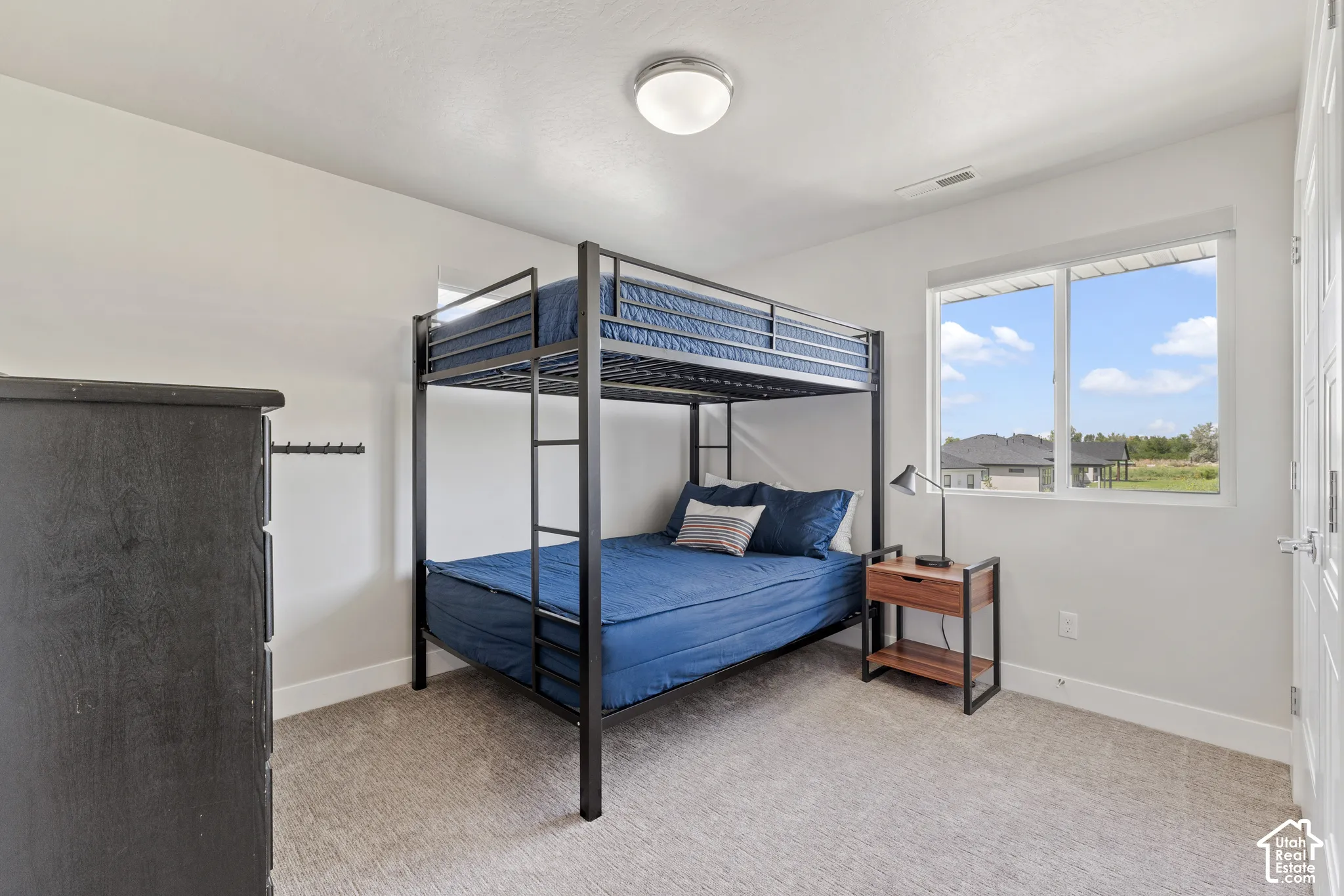 Bright secondary bedroom featuring a metal canopy bed frame, carpeted flooring, and abundant natural light from large windows overlooking the surrounding landscape