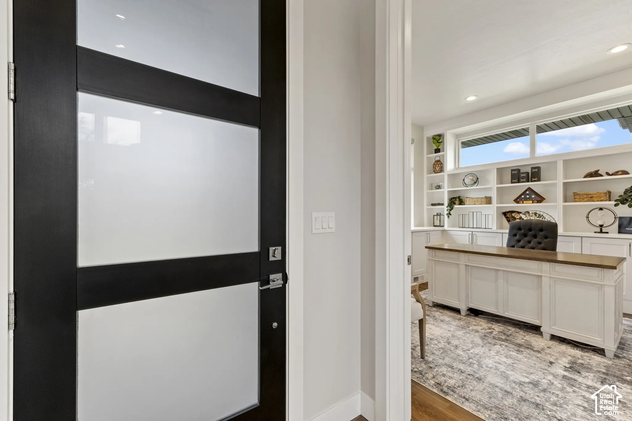Modern interior entry featuring a contemporary black-framed glass and white panel door with integrated smart lock hardware, opening to a bright home office with built-in white cabinetry, wood-finish countertops, and expansive windows providing natural lig